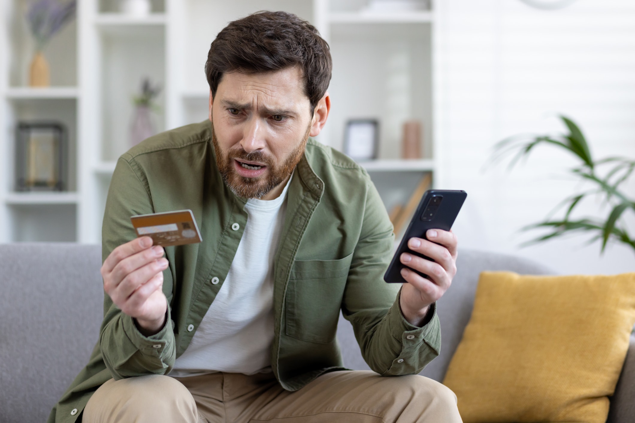 upset cheated man sitting on couch in living room at home, holding bank credit card and phone, rejected money transfer, negative balance, scam money loss