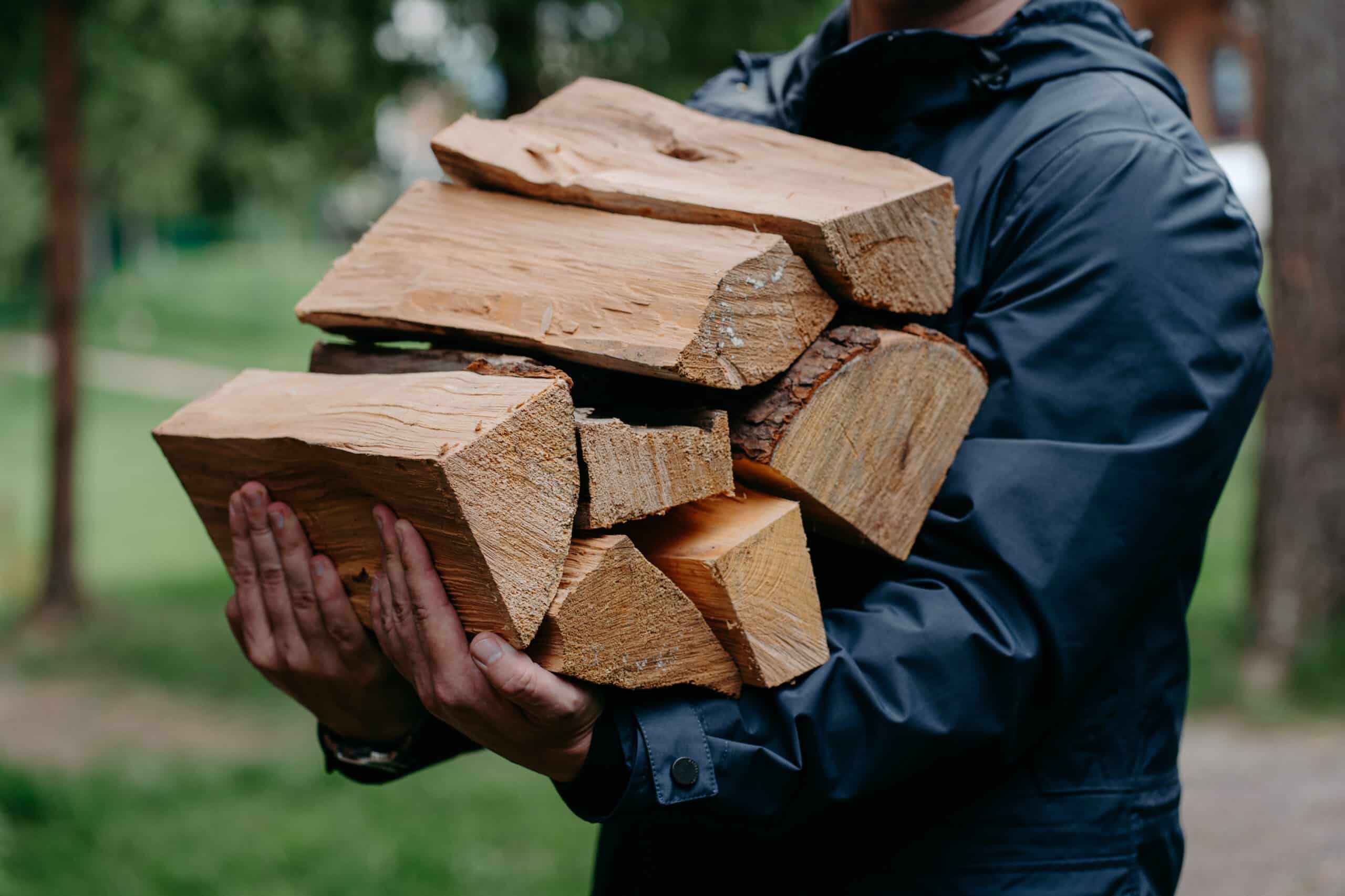 cropped shot of faceless man in black jacket carries pile of firewood poses against blurred forest background.